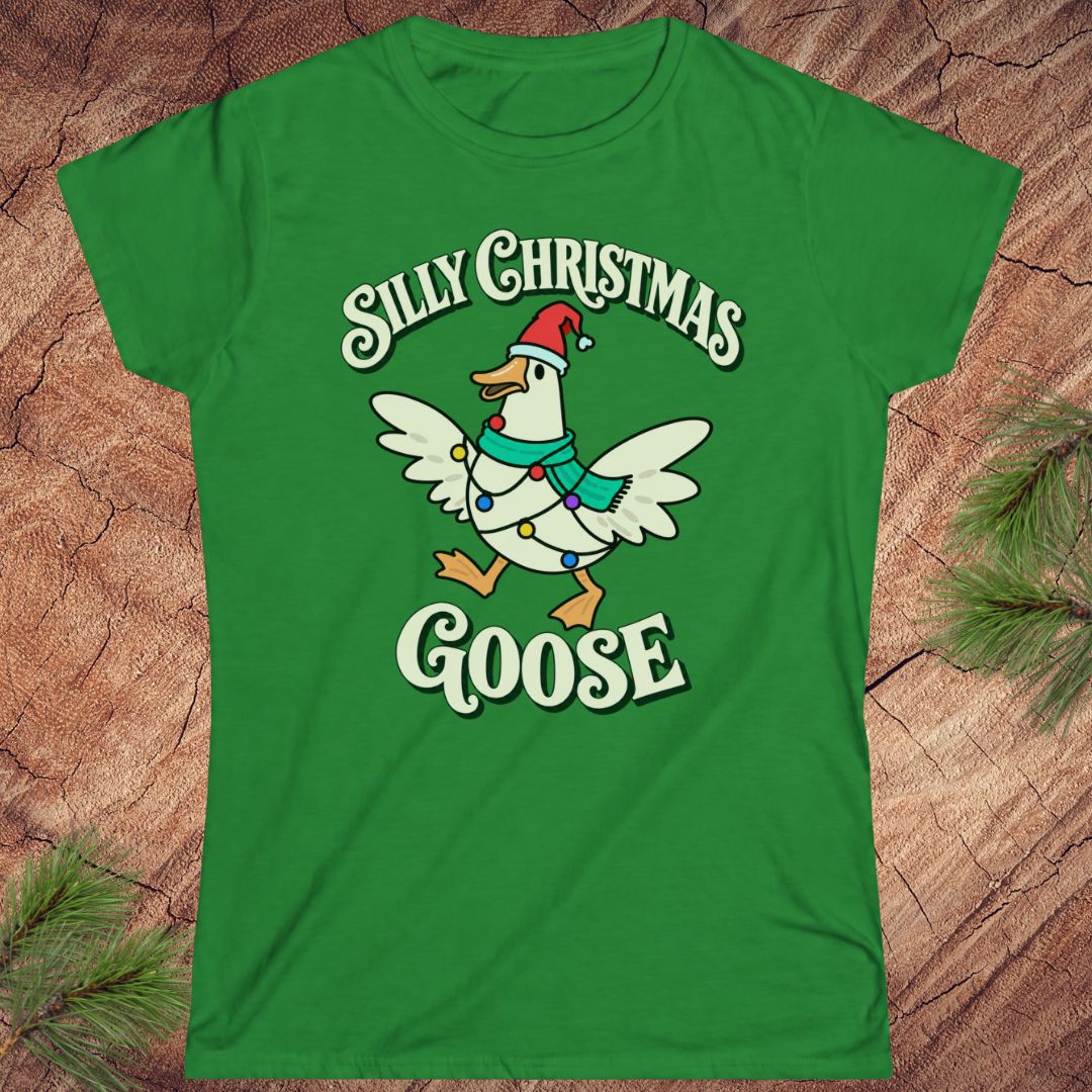 Irish green women's t-shirt with a goose in a Santa hat and teal scarf, wrapped in colorful Christmas lights under retro Silly Christmas Goose lettering, laid flat on a rustic wood background with pine branches.