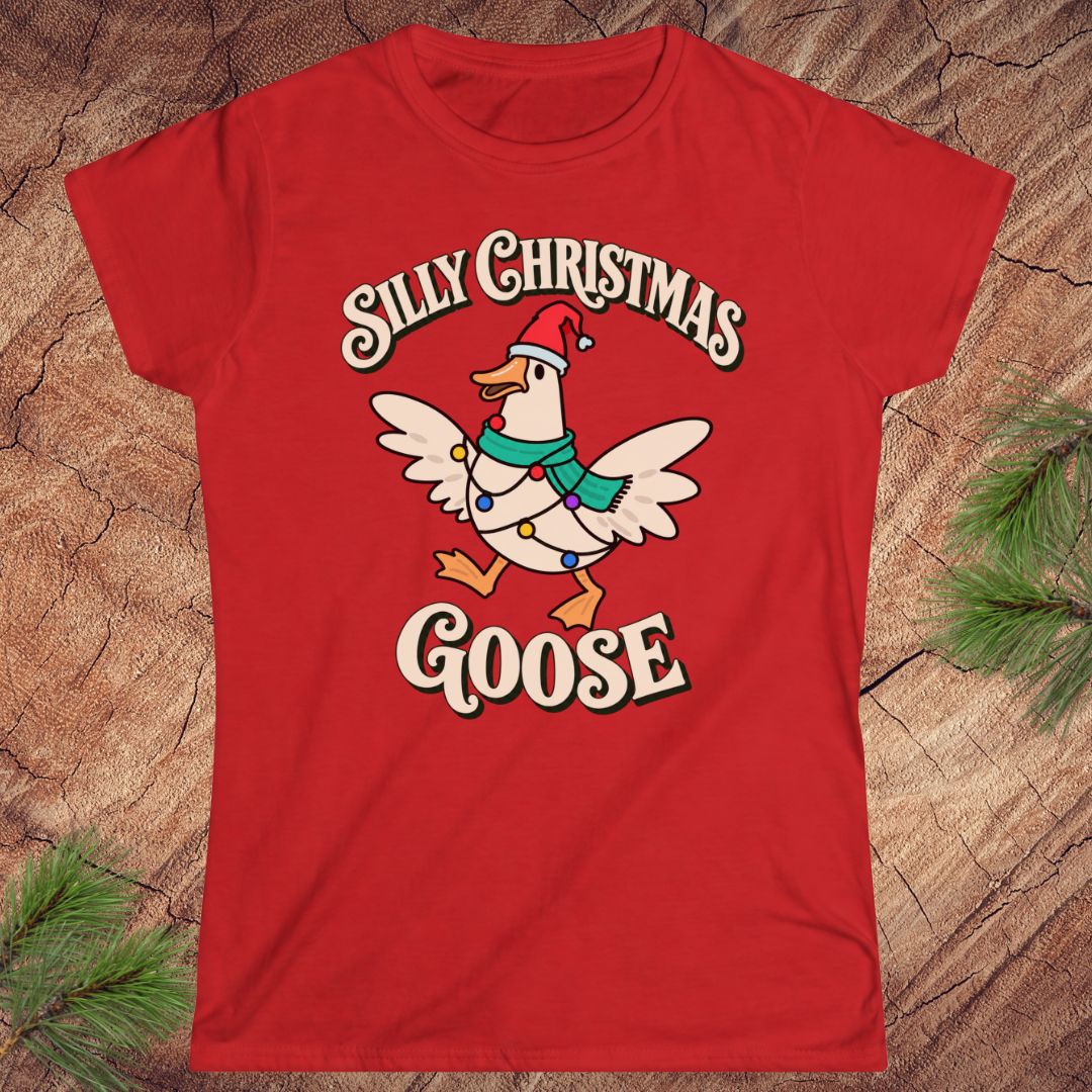 Red women's t-shirt with a goose in a Santa hat and teal scarf, wrapped in colorful Christmas lights under retro Silly Christmas Goose lettering, laid flat on a rustic wood background with pine branches.