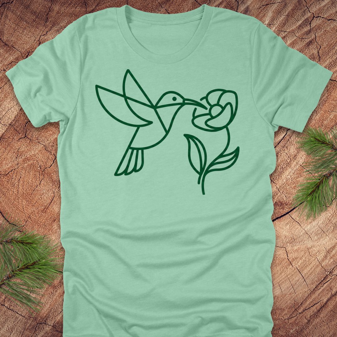 Light green t-shirt with a black line drawing of a hummingbird and flower on a wooden surface.