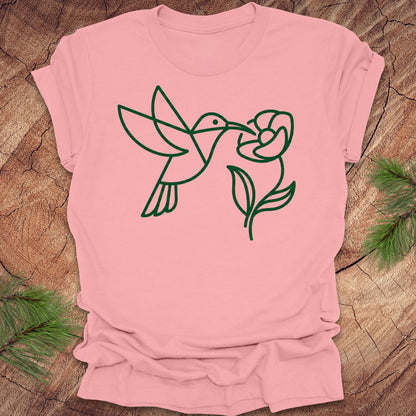 Pink t-shirt with green line art of a hummingbird and flower on a wooden surface.