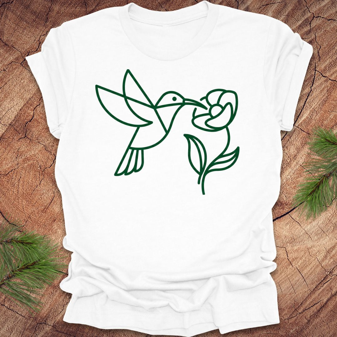 White t-shirt with green line art of a hummingbird and flower on a wooden surface.