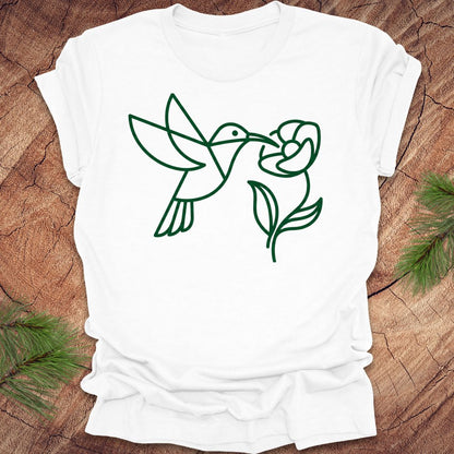 White t-shirt with green line art of a hummingbird and flower on a wooden surface.