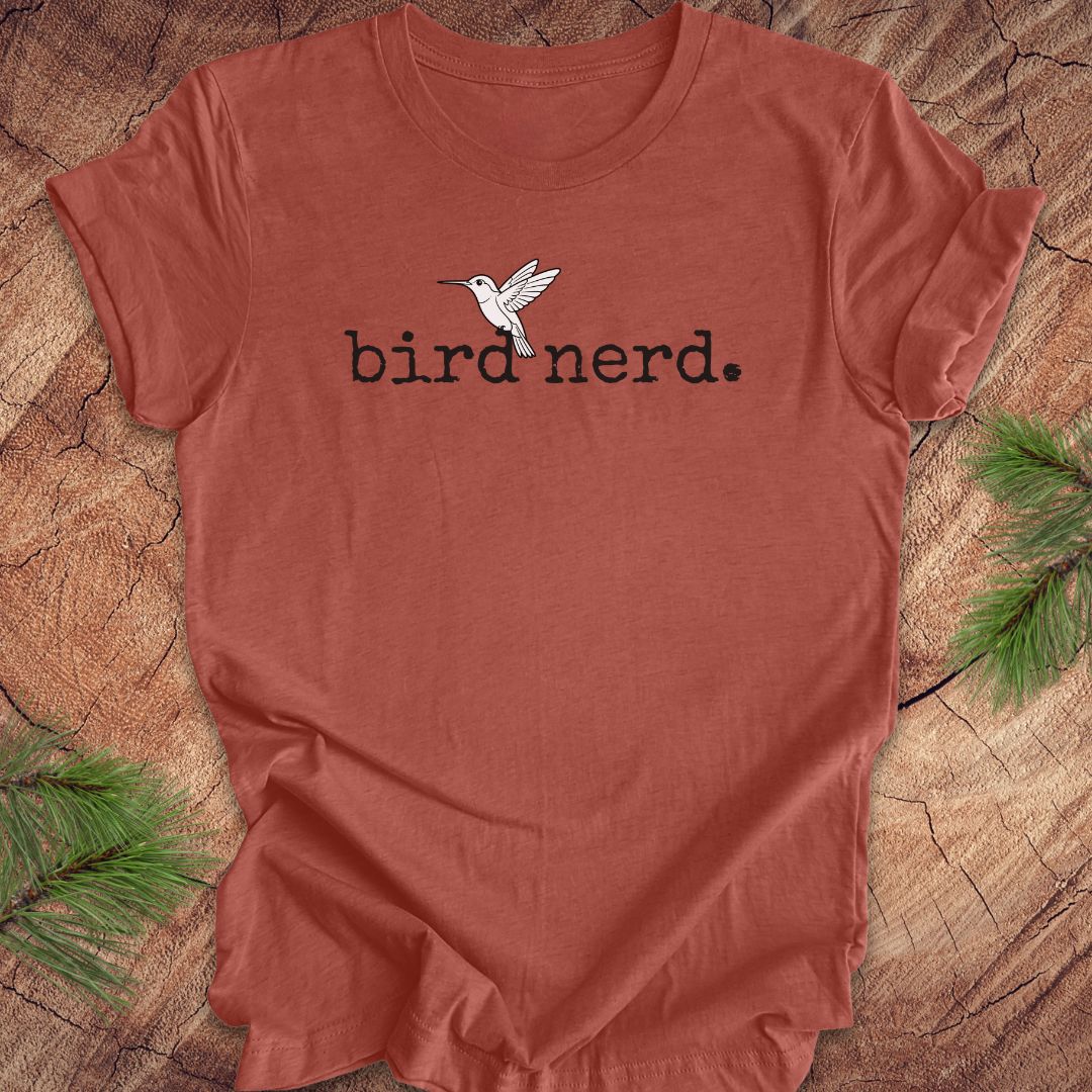 A terracotta-colored short-sleeve t-shirt with a black 'bird nerd.' text graphic and a white hummingbird illustration on the chest, laid flat on a rustic wood slice background with pine branches in the corners.