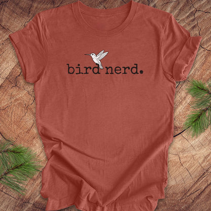 A terracotta-colored short-sleeve t-shirt with a black 'bird nerd.' text graphic and a white hummingbird illustration on the chest, laid flat on a rustic wood slice background with pine branches in the corners.