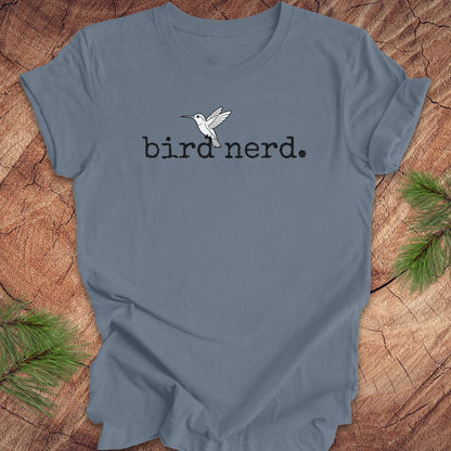 A slate blue crewneck t-shirt featuring a black printed text 'bird nerd.' and a white hummingbird graphic on the chest, displayed flat on a rustic wood background with pine needles.