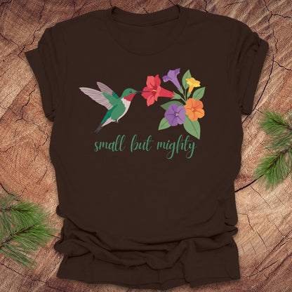 Brown t-shirt with a hummingbird and flowers with text, 'small but mighty' on a wooden surface