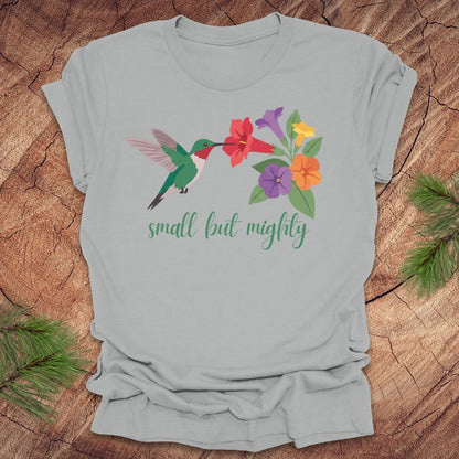 Silver t-shirt with a hummingbird and flowers with text, 'small but mighty' on a wooden surface