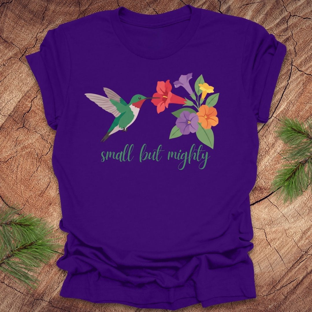 Purple t-shirt with a hummingbird and flowers with text, 'small but mighty' on a wooden surface