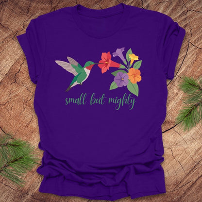 Purple t-shirt with a hummingbird and flowers with text, 'small but mighty' on a wooden surface