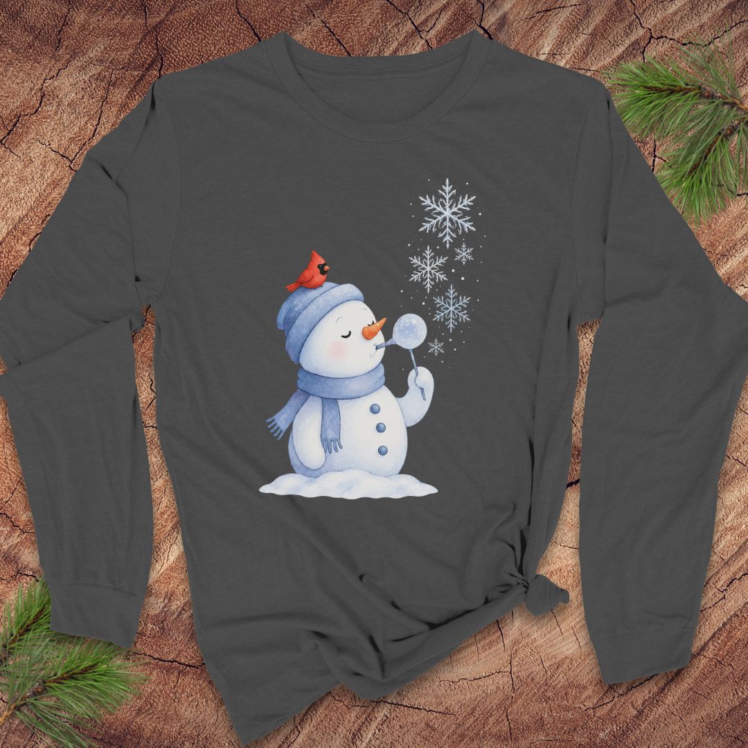 A dark gray long-sleeve t-shirt with a winter snowman graphic, laid flat on a rustic wood background with pine branches in the corners.