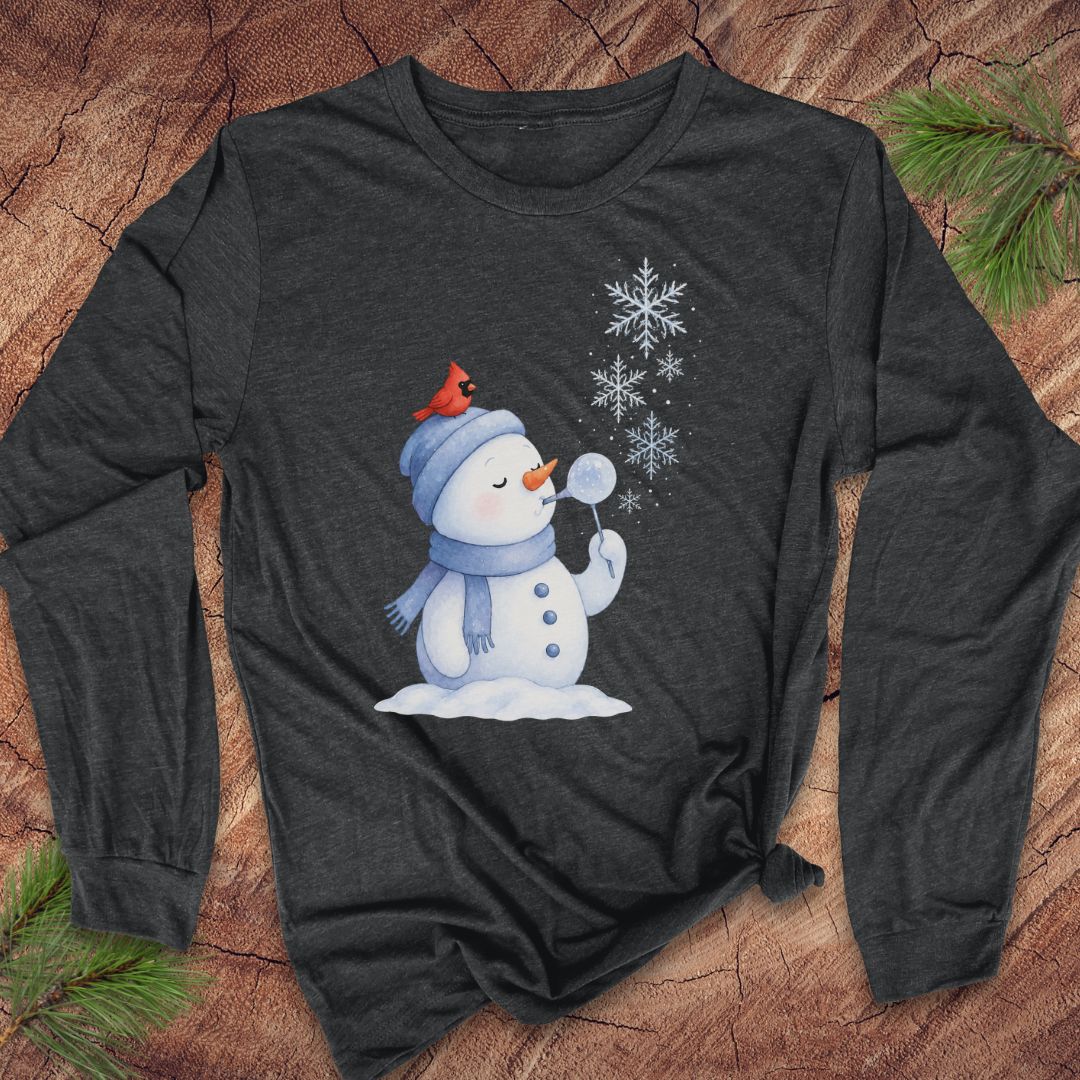 A dark charcoal gray long-sleeve t-shirt with a winter holiday graphic of a snowman blowing snowflakes, styled flat on a rustic wood background with pine needles.