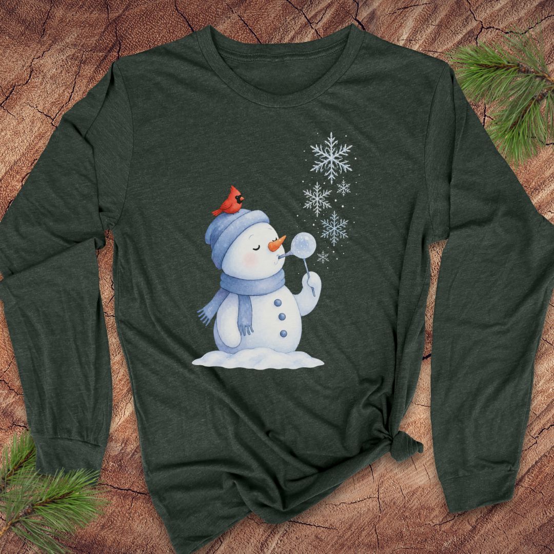 A dark heather green long-sleeve t-shirt with a winter graphic of a snowman blowing snowflakes, featuring a small red cardinal on his hat. The shirt is laid flat on a rustic wood background with pine branches in the corners.