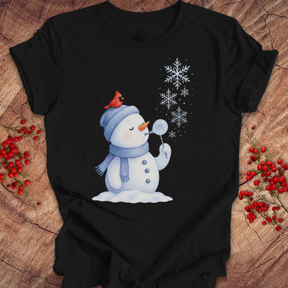 A black short-sleeve t-shirt with a winter holiday graphic of a snowman blowing snow, styled flat on a rustic wood background with red berries.