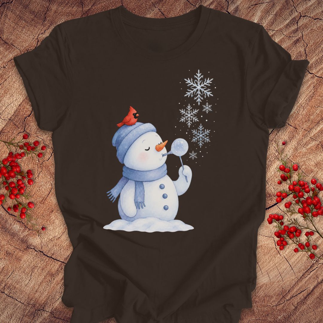 A dark brown short-sleeve t-shirt with a winter graphic of a snowman blowing snowflakes, featuring a red cardinal on its hat, laid flat on a rustic wood background with red berries.