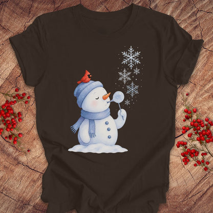 A dark brown short-sleeve t-shirt with a winter graphic of a snowman blowing snowflakes, featuring a red cardinal on its hat, laid flat on a rustic wood background with red berries.