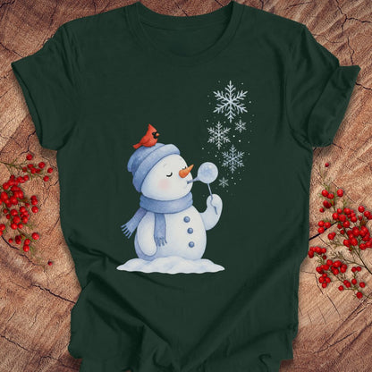 A dark forest green short-sleeve t-shirt with a winter graphic of a snowman blowing snowflakes, styled flat on a rustic wood background with red berries.