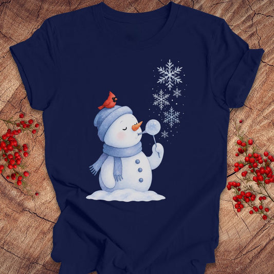 A navy blue short-sleeve t-shirt with a winter holiday graphic of a snowman blowing a bubble with a red cardinal on his hat, displayed on a rustic wood background with red berries.