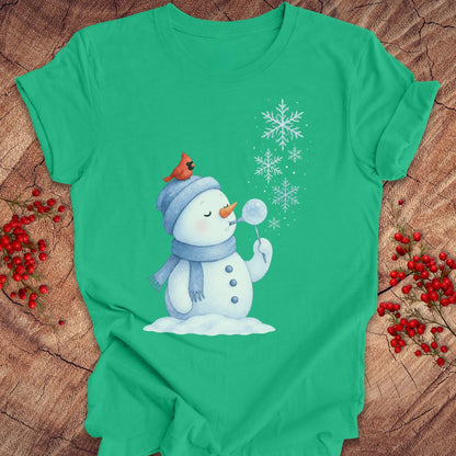 A green short-sleeve t-shirt with a winter holiday graphic of a snowman blowing a snowball, with a small red cardinal on its hat. The shirt is laid flat on a rustic wood background decorated with red berries.