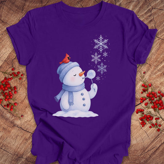 A purple short-sleeve t-shirt with a cartoon snowman graphic, laid flat on a rustic wood background with red berries.