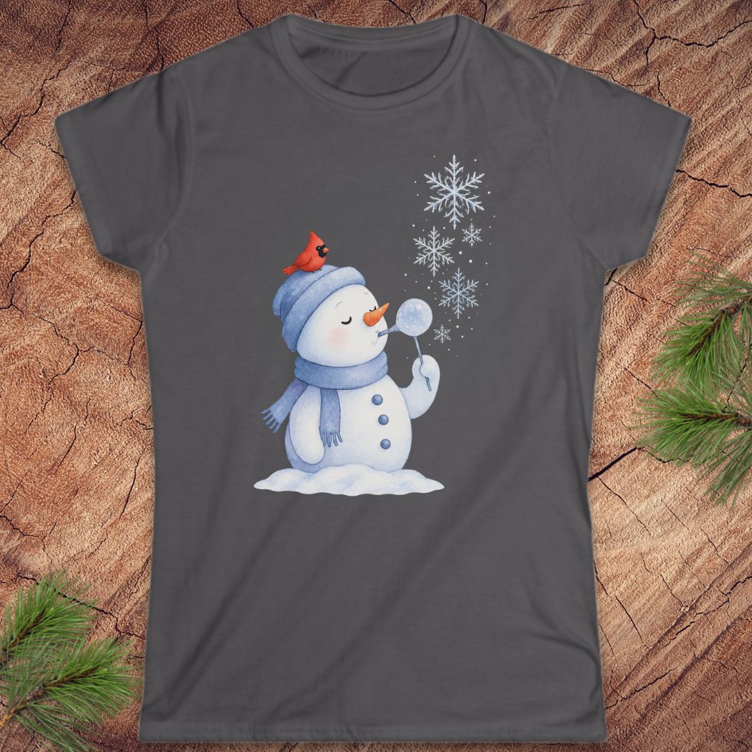 A dark gray short-sleeve crewneck t-shirt featuring a festive graphic of a snowman wearing a blue beanie and scarf, with a small red cardinal perched on the beanie. The snowman blows white snowflakes, with several large snowflakes floating above.