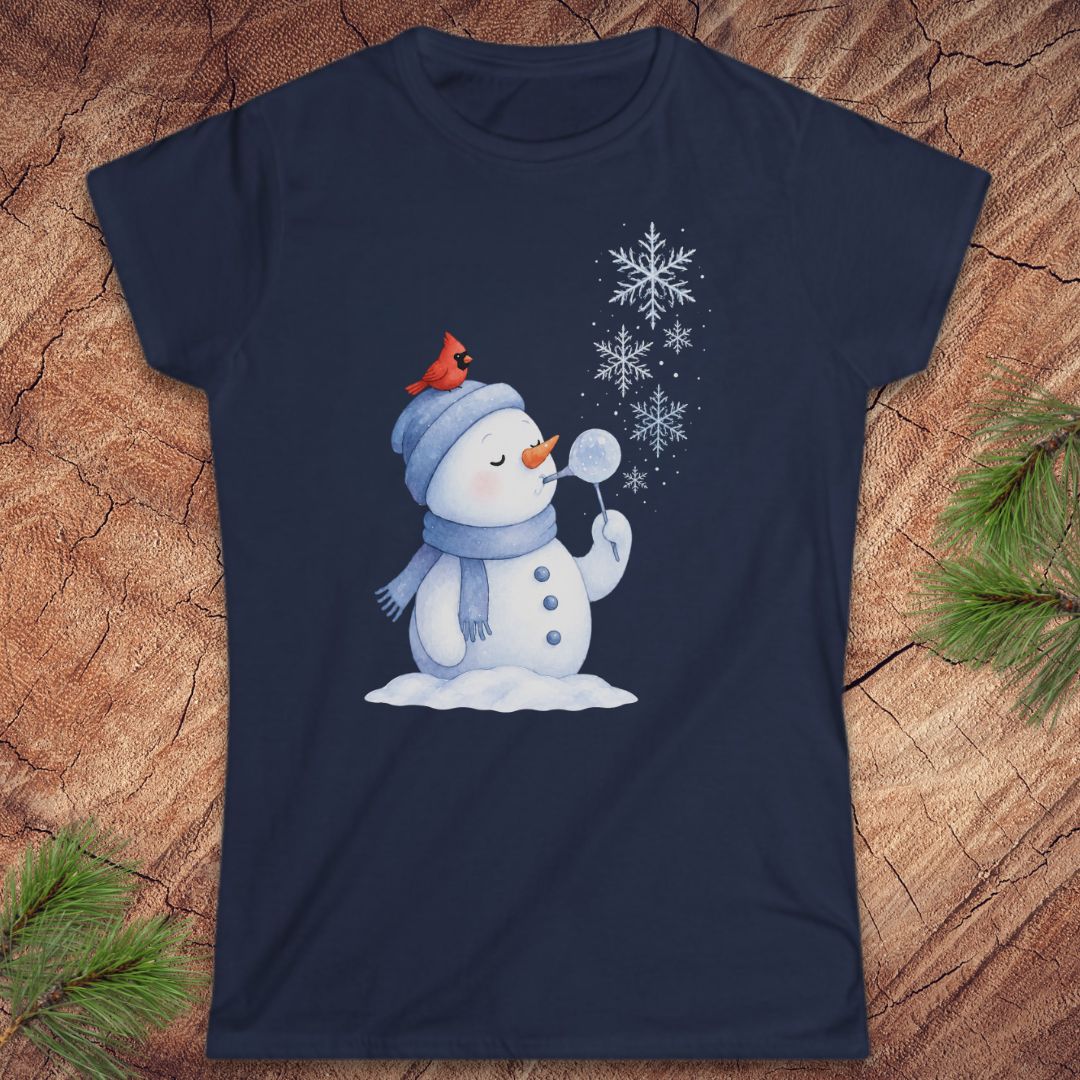 A navy blue short-sleeve t-shirt with a winter graphic of a snowman wearing a red cardinal hat, surrounded by snowflakes, displayed on a rustic wood background with pine branches.