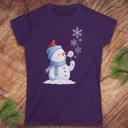 A purple short-sleeve crewneck t-shirt featuring a cartoon snowman graphic with a red cardinal on its hat, surrounded by white snowflakes. The t-shirt is displayed flat on a textured wood background with pine branches in the corners.