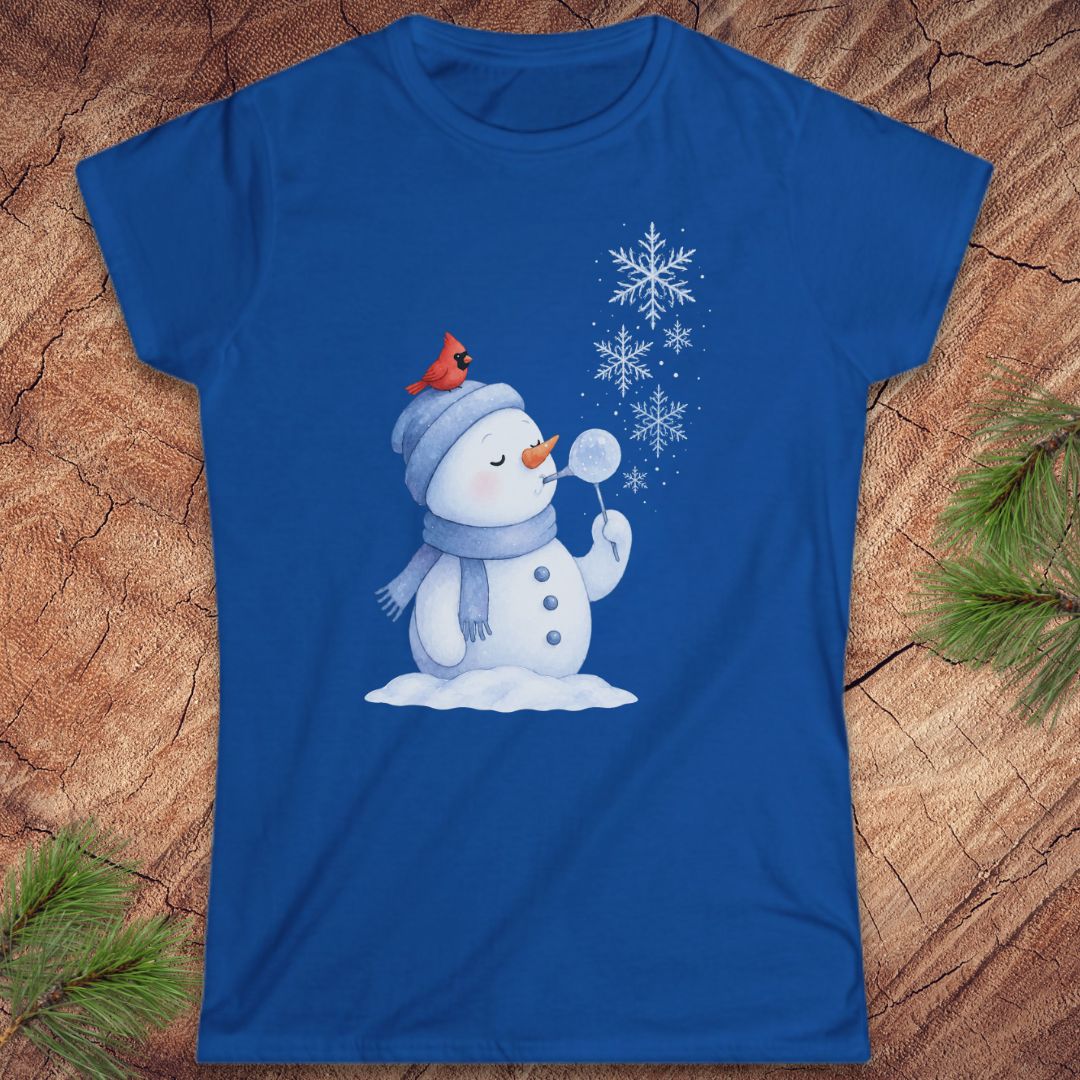 A royal blue short-sleeve t-shirt with a winter snowman graphic, laid flat on a textured wood background with pine branches in the corners. The snowman is wearing a gray beanie with a small red cardinal on top, blowing white snowflakes.