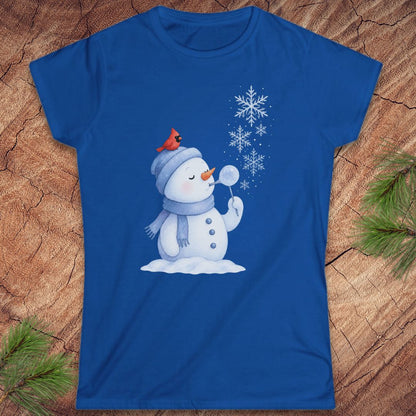 A royal blue short-sleeve t-shirt with a winter snowman graphic, laid flat on a textured wood background with pine branches in the corners. The snowman is wearing a gray beanie with a small red cardinal on top, blowing white snowflakes.