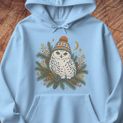 Light blue hoodie with a Snowy Owl in an orange pom-pom knit beanie, nestled in pine boughs, pinecones, and red berries under a crescent moon, laid flat on a rustic wood background.