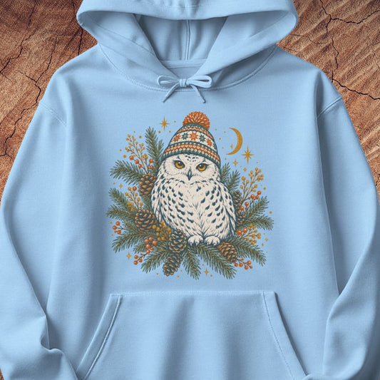 Light blue hoodie with a Snowy Owl in an orange pom-pom knit beanie, nestled in pine boughs, pinecones, and red berries under a crescent moon, laid flat on a rustic wood background.
