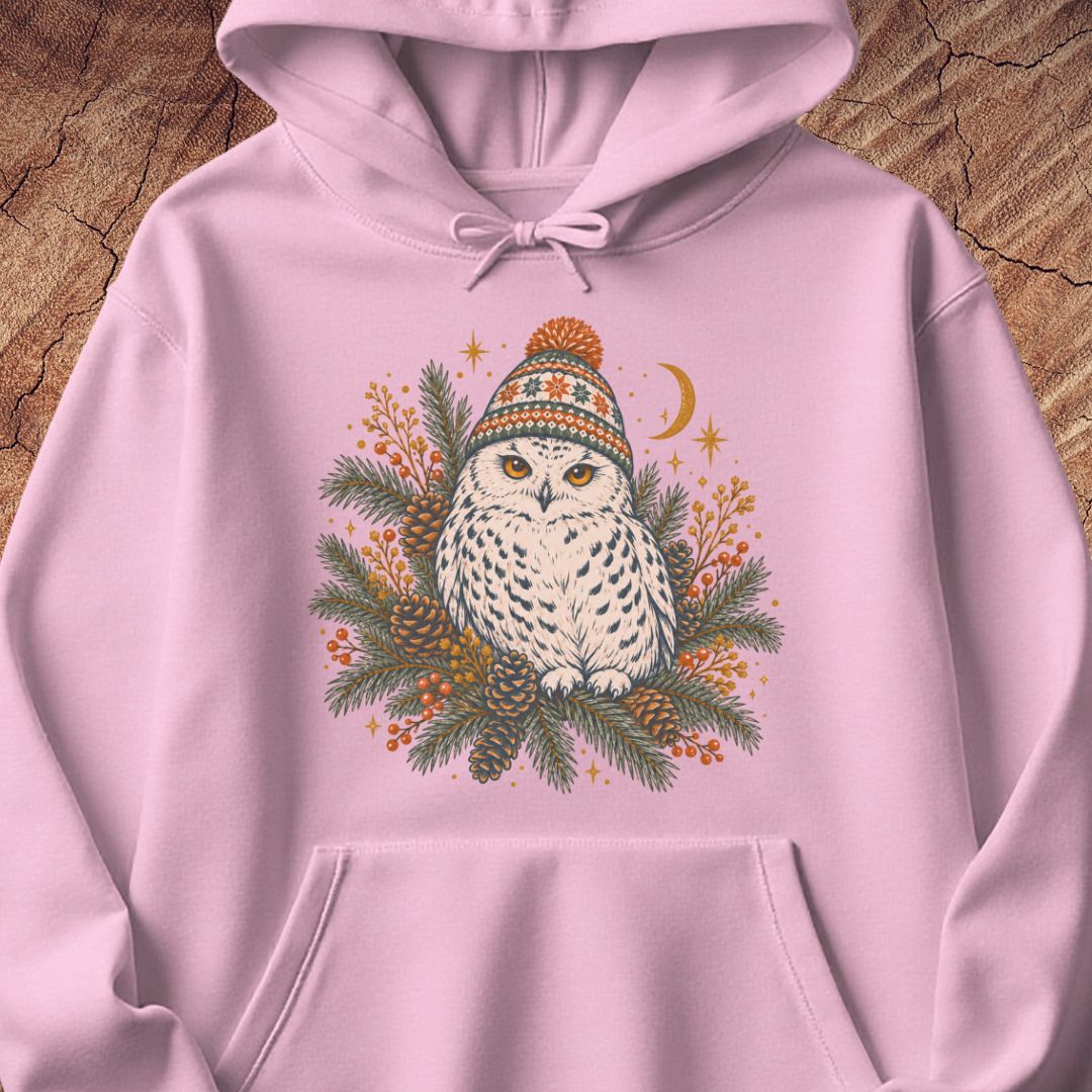 Light pink hoodie with a Snowy Owl in an orange pom-pom knit beanie, nestled in pine boughs, pinecones, and red berries under a crescent moon, laid flat on a rustic wood background.