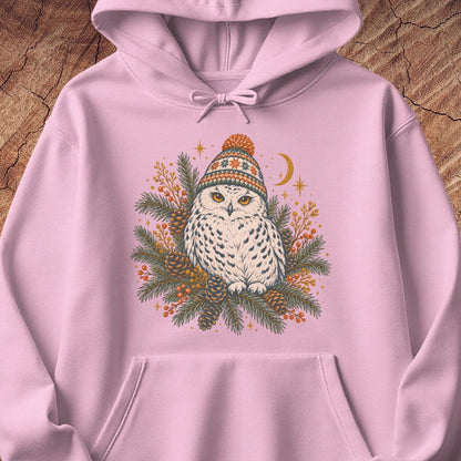 Light pink hoodie with a Snowy Owl in an orange pom-pom knit beanie, nestled in pine boughs, pinecones, and red berries under a crescent moon, laid flat on a rustic wood background.