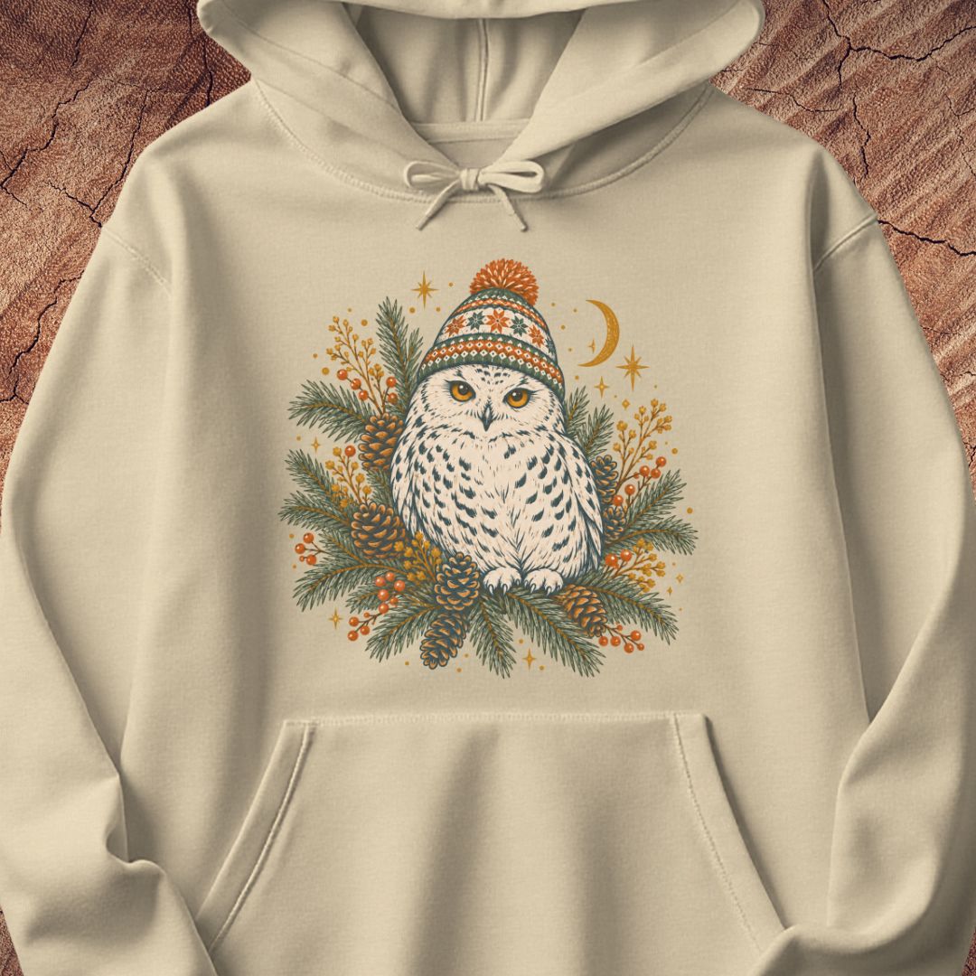 Sand beige hoodie with a Snowy Owl in an orange pom-pom knit beanie, nestled in pine boughs, pinecones, and red berries under a crescent moon, laid flat on a rustic wood background.
