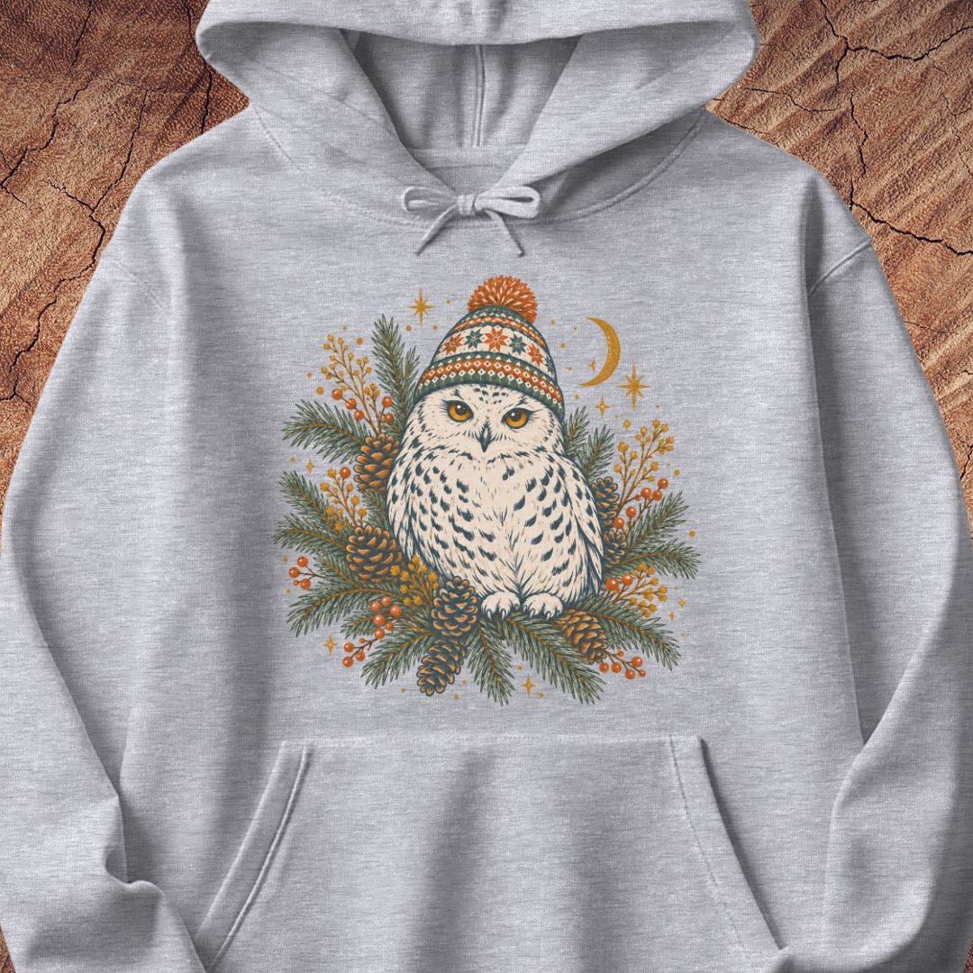 Sport grey hoodie with a Snowy Owl in an orange pom-pom knit beanie, nestled in pine boughs, pinecones, and red berries under a crescent moon, laid flat on a rustic wood background.