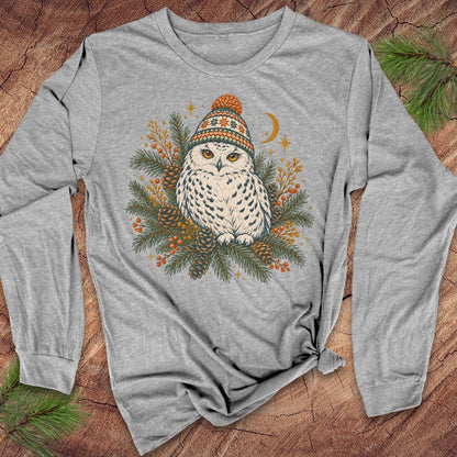 Athletic heather gray long-sleeve t-shirt with a Snowy Owl in an orange pom-pom knit beanie, nestled in pine boughs, pinecones, and red berries under a crescent moon, laid flat on a rustic wood background with pine branches.