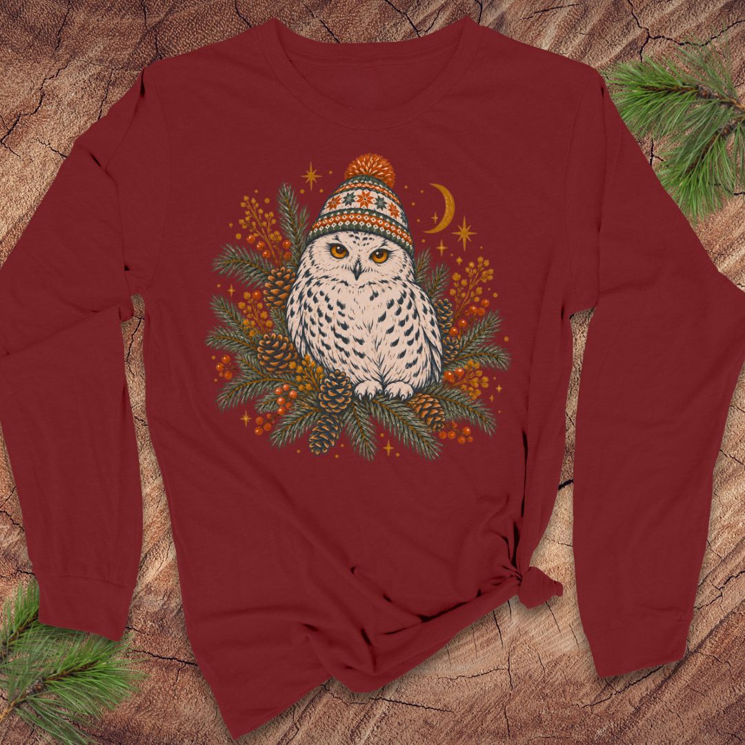 Cardinal red long-sleeve t-shirt with a Snowy Owl in an orange pom-pom knit beanie, nestled in pine boughs, pinecones, and red berries under a crescent moon, laid flat on a rustic wood background with pine branches.