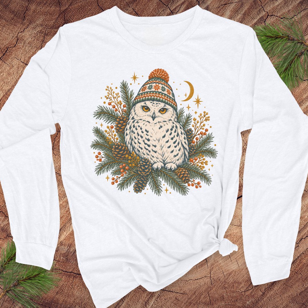 White long-sleeve t-shirt with a Snowy Owl in an orange pom-pom knit beanie, nestled in pine boughs, pinecones, and red berries under a crescent moon, laid flat on a rustic wood background with pine branches.