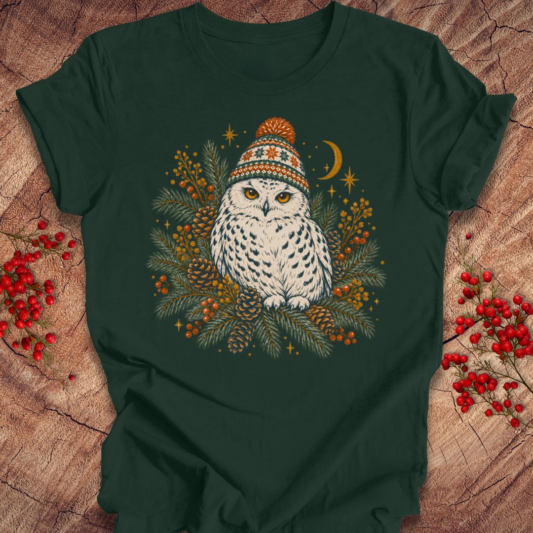 A dark forest green t-shirt laid flat on a rustic wood background, featuring a central graphic of a white owl wearing a colorful knit winter hat, surrounded by pine branches, berries, pinecones, and a crescent moon.