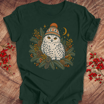 A dark forest green t-shirt laid flat on a rustic wood background, featuring a central graphic of a white owl wearing a colorful knit winter hat, surrounded by pine branches, berries, pinecones, and a crescent moon.