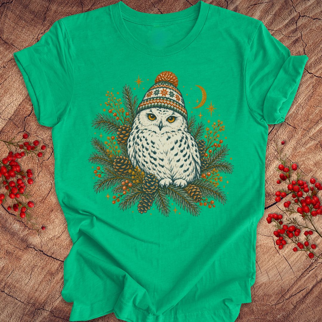 A bright green short-sleeve t-shirt featuring a graphic of a snowy owl wearing a colorful knit hat, surrounded by pine branches, pinecones, and red berries, laid flat on a rustic wood background.
