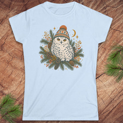 Light blue women's t-shirt with a Snowy Owl in an orange pom-pom knit beanie, nestled in pine boughs, pinecones, and red berries under a crescent moon, laid flat on a rustic wood background with pine branches.