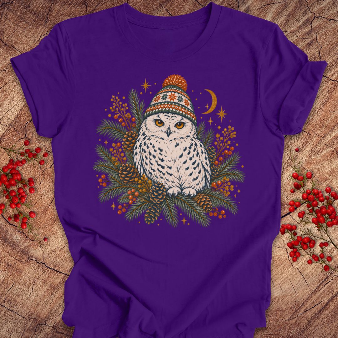 A purple short-sleeve crewneck t-shirt featuring a graphic of a snowy owl wearing a colorful knit hat, surrounded by pine branches, berries, stars, and a crescent moon, laid flat on a rustic wood background with red berries.