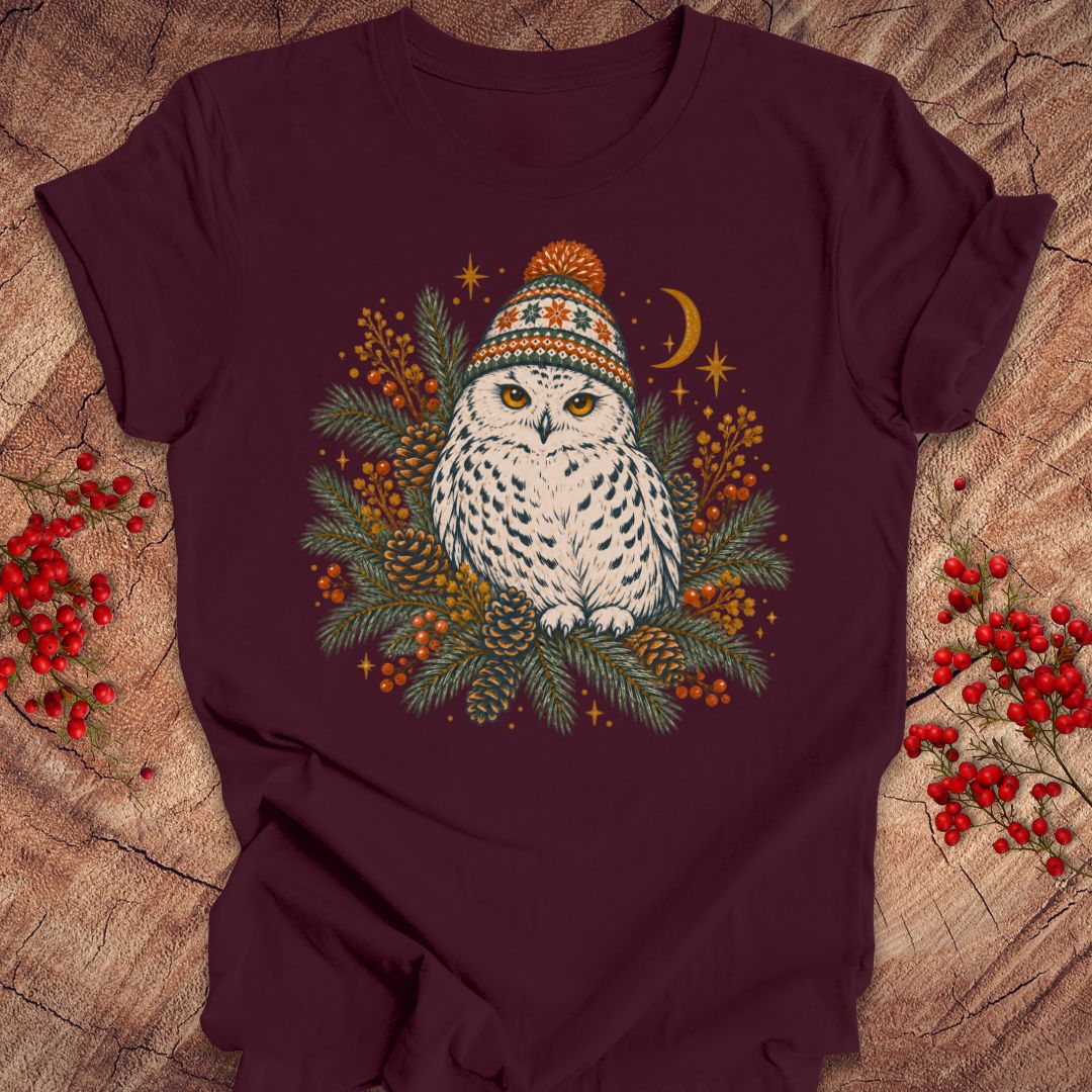 A dark maroon short-sleeve t-shirt laid flat on a rustic wood surface, featuring a central graphic of a white owl wearing a colorful knit hat surrounded by green pine branches, orange berries, stars, and a crescent moon. Red berries are placed decoratively on both sides of the shirt.