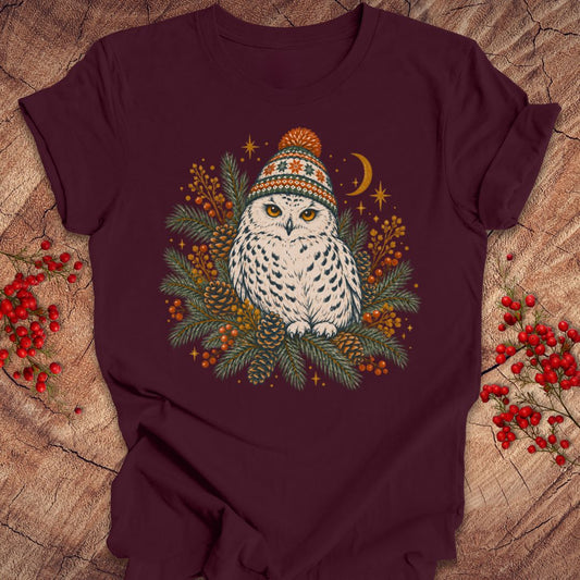 A dark maroon short-sleeve t-shirt laid flat on a rustic wood surface, featuring a central graphic of a white owl wearing a colorful knit hat surrounded by green pine branches, orange berries, stars, and a crescent moon. Red berries are placed decoratively on both sides of the shirt.