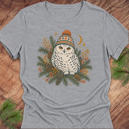 Athletic heather gray women's relaxed t-shirt with a Snowy Owl in an orange pom-pom knit beanie, nestled in pine boughs, pinecones, and red berries under a crescent moon, laid flat on a rustic wood background with pine branches.