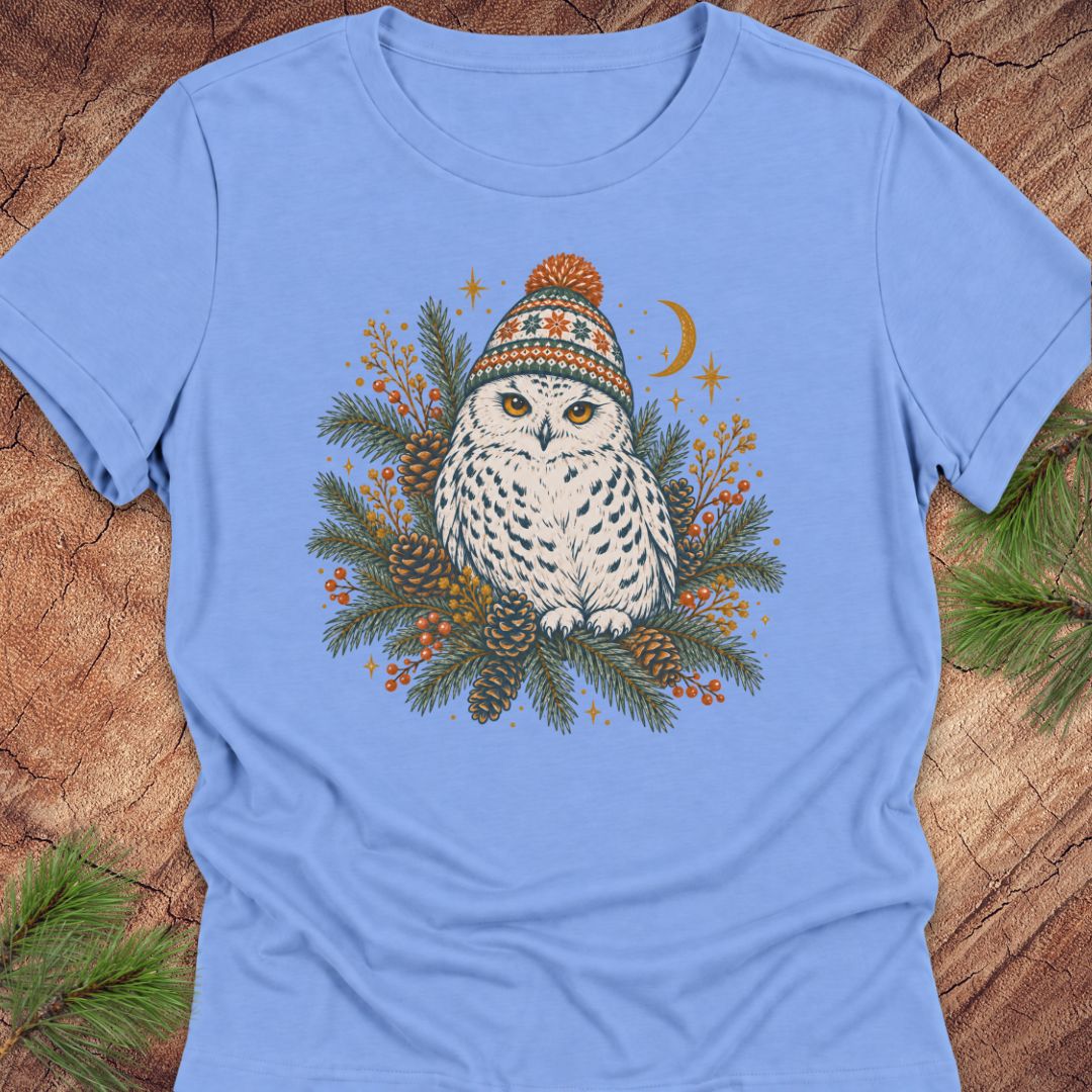 Carolina blue women's relaxed t-shirt with a Snowy Owl in an orange pom-pom knit beanie, nestled in pine boughs, pinecones, and red berries under a crescent moon, laid flat on a rustic wood background with pine branches.