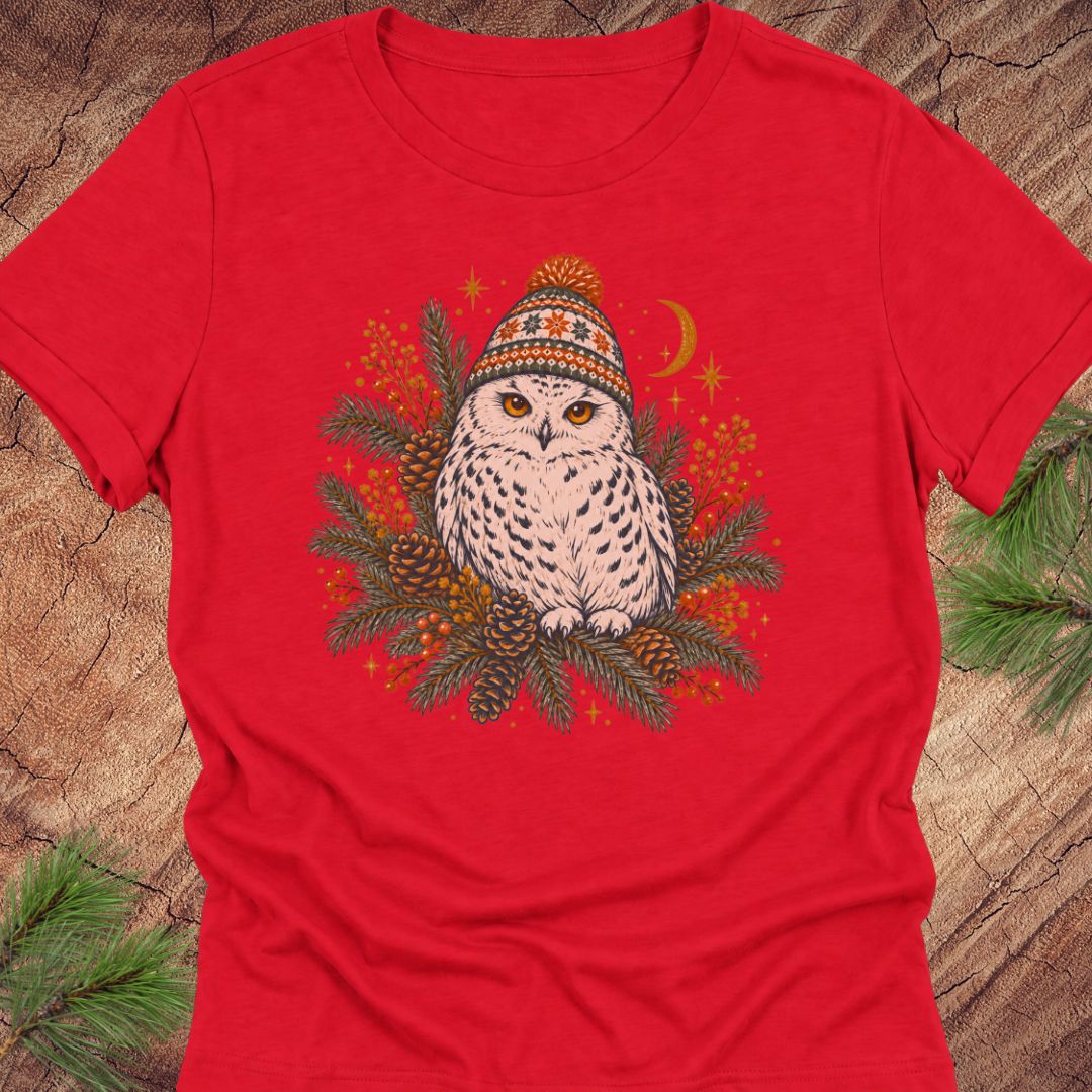 Red women's relaxed t-shirt with a Snowy Owl in an orange pom-pom knit beanie, nestled in pine boughs, pinecones, and red berries under a crescent moon, laid flat on a rustic wood background with pine branches.
