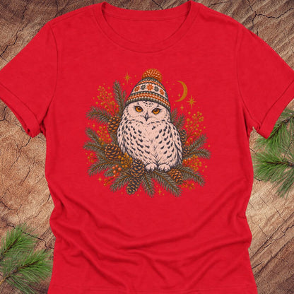 Red women's relaxed t-shirt with a Snowy Owl in an orange pom-pom knit beanie, nestled in pine boughs, pinecones, and red berries under a crescent moon, laid flat on a rustic wood background with pine branches.
