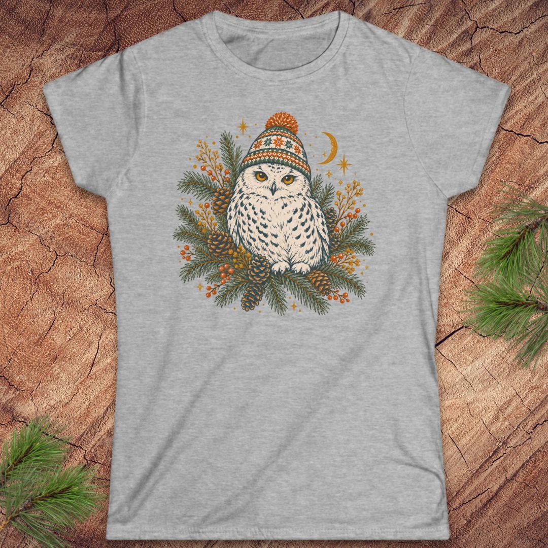 Sport grey women's t-shirt with a Snowy Owl in an orange pom-pom knit beanie, nestled in pine boughs, pinecones, and red berries under a crescent moon, laid flat on a rustic wood background with pine branches.