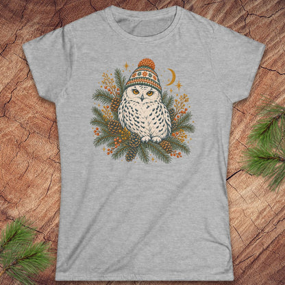 Sport grey women's t-shirt with a Snowy Owl in an orange pom-pom knit beanie, nestled in pine boughs, pinecones, and red berries under a crescent moon, laid flat on a rustic wood background with pine branches.